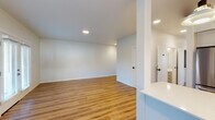 This contemporary single-floor unit showcases a thoughtfully designed open-concept layout with a cohesive, modern aesthetic throughout. Light wood luxury vinyl plank flooring runs seamlessly across the entire space, complementing the crisp white walls and creating a bright, airy atmosphere. Recessed lighting provides consistent illumination throughout the living areas.

The kitchen is a standout feature, equipped with white flat-front cabinetry, quartz countertops, a subway tile backsplash, and a matte black faucet over a stainless steel sink. A pair of natural live-edge wood open shelves add warmth and character against the otherwise clean design. A peninsula with an overhang provides additional counter space and separates the kitchen from the dining area, which benefits from a large window with horizontal blinds allowing natural light to filter in.

The spacious living area flows naturally from the kitchen and dining zones, with a sliding glass door that leads to an exterior access point, further enhancing natural light. The bedrooms are well-proportioned, each featuring windows overlooking the surrounding neighborhood and the same consistent wood flooring.

Both bathrooms maintain the modern design language, outfitted with white vanities, quartz countertops, large mirrors, matte black hardware and fixtures, and tub-shower combinations — all set against clean white walls and matching wood-tone flooring.