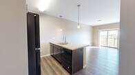 Located at 1602 Belsly Blvd in Moorhead, MN, this one-bedroom, one-bathroom unit offers a well-designed, single-floor layout that maximizes every inch of its compact footprint. The open-concept kitchen and living area serves as the heart of the unit, featuring rich dark cabinetry, granite-style countertops, a peninsula with an undermount sink, and black appliances including a refrigerator, electric range, microwave, and dishwasher. Pendant lighting over the peninsula adds a modern touch, while hardwood-style flooring runs throughout the main living areas, lending warmth and continuity. A sliding glass door in the living area opens to a private balcony, offering outdoor views of the surrounding Moorhead neighborhood and filling the space with natural light. The bedroom is carpeted for comfort and includes a window with blinds, a wall-mounted AC unit, and baseboard heating. It connects directly to the full bathroom, which features tile flooring, a granite-topped dark vanity, and a full tub-shower combination. A dedicated in-unit laundry area includes wire shelving for additional storage. Neutral warm-beige tones throughout create a cohesive and inviting atmosphere.