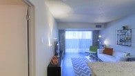 This single-floor space offers a well-designed, open-concept layout that maximizes functionality and comfort. The living area is the centerpiece of the home, anchored by large sliding glass doors fitted with vertical blinds and bold geometric-patterned curtains, allowing abundant natural light to flow throughout the interior. The hardwood-style flooring adds warmth and a contemporary feel to the space, complementing the neutral-toned walls with their subtle textured finish. A textured popcorn ceiling adds a classic architectural touch overhead, while recessed HVAC vents ensure efficient climate control. The living area is well-proportioned, offering ample room for seating and entertainment furniture. A sleek, low-profile media console anchors one side of the room, while the seating arrangement — including a modern accent chair in a vibrant green — introduces a lively pop of color against the otherwise neutral palette. Decorative elements such as a sculptural orb accent piece and tall floor lamps contribute to the room's curated, stylish ambiance. A granite-surfaced counter or bar edge is visible at the edge of the frame, hinting at an adjacent kitchen or dining area. Overall, the space presents a clean, modern aesthetic with a welcoming atmosphere enhanced by thoughtful décor and generous natural light.