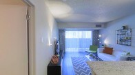 This single-floor space offers a well-designed, open-concept layout that maximizes functionality and comfort. The living area is the centerpiece of the home, anchored by large sliding glass doors fitted with vertical blinds and bold geometric-patterned curtains, allowing abundant natural light to flow throughout the interior. The hardwood-style flooring adds warmth and a contemporary feel to the space, complementing the neutral-toned walls with their subtle textured finish. A textured popcorn ceiling adds a classic architectural touch overhead, while recessed HVAC vents ensure efficient climate control. The living area is well-proportioned, offering ample room for seating and entertainment furniture. A sleek, low-profile media console anchors one side of the room, while the seating arrangement — including a modern accent chair in a vibrant green — introduces a lively pop of color against the otherwise neutral palette. Decorative elements such as a sculptural orb accent piece and tall floor lamps contribute to the room's curated, stylish ambiance. A granite-surfaced counter or bar edge is visible at the edge of the frame, hinting at an adjacent kitchen or dining area. Overall, the space presents a clean, modern aesthetic with a welcoming atmosphere enhanced by thoughtful décor and generous natural light.