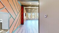 Located at 2025 Iris Dr SE in Conyers, Georgia, this single-floor open-plan studio is a visually dynamic and well-appointed space designed for activity and versatility. The interior immediately draws the eye with a striking feature wall finished in a bold orange geometric chevron pattern, accented by teal trim lines — a design choice that energizes the entire room and establishes a distinct, modern aesthetic.

The ceiling is one of the space's most notable architectural elements, showcasing exposed wooden planks set between structural steel beams with recessed lighting integrated throughout, creating a warm industrial character. The flooring transitions between smooth hardwood-style planks and a finished concrete mat area, suggesting a dual-purpose layout suited for both active and flexible use.

Natural light pours in through multiple large, black-framed windows along the exterior walls, offering views of the surrounding outdoor area, including a pool and landscaped grounds. This abundance of glazing gives the space an open, airy feel throughout the day.

A kitchenette or wet bar area is visible through a passage in the feature wall, adding functional convenience. An exterior door provides direct outdoor access, reinforcing the space's connection to the surrounding amenities. Overall, the studio offers a bold, purpose-built environment ideal for fitness, wellness, or community programming.