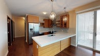 This single-floor space is anchored by a thoughtfully designed modern kitchen that serves as its centerpiece. The kitchen showcases warm, medium-toned wood cabinetry that lines the walls, offering generous storage alongside open glass-front upper shelving for display. A standout feature is the expansive kitchen island, finished with a crisp white countertop and a built-in sink, providing ample workspace and a natural gathering point. Overhead, two striking spherical globe pendant lights suspended from the ceiling add a refined, contemporary touch that elevates the overall aesthetic. The cooking area is equipped with stainless steel appliances, including a French door refrigerator with an in-door water and ice dispenser, a range, and a ventilation hood, all set against a subtly textured tile backsplash that adds depth and visual interest. Rich, dark hardwood flooring runs throughout, creating a warm contrast to the lighter cabinetry and countertops. Natural light filters in through window treatments, enhancing the bright and welcoming atmosphere. The overall design balances functionality with modern style, resulting in a polished, cohesive living space.