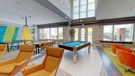 This single-floor community amenity space is designed with a contemporary aesthetic and a lively, social atmosphere. The main gathering area commands attention with soaring double-height ceilings, large floor-to-ceiling windows, and an abundance of natural light. A sleek pool table with chrome legs and a teal felt surface serves as the centerpiece, surrounded by comfortable tan leather lounge chairs arranged for easy conversation and casual gatherings.

The fully equipped kitchen features crisp white cabinetry, dark granite countertops, a textured metallic accent wall, a wall-mounted flat-screen TV, a built-in wine cooler, and a spacious central island with a white surface and vibrant orange bar stools — ideal for group entertaining or informal dining.

A cozy lounge nook provides a relaxed retreat, with a long built-in leather bench accented by patterned cushions, wall sconces, and bright teal accent walls. Large windows frame views of the surrounding landscape, filling the space with warmth and natural light.

A game table area with a chess set sits adjacent to full-height folding glass doors that open directly onto a covered outdoor patio, complete with dining seating, ceiling fans, and an outdoor grill. This seamless indoor-outdoor connection enhances the overall versatility and appeal of the space.