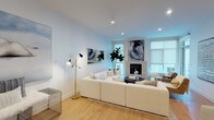 This single-floor residence showcases a thoughtfully designed open-concept layout with a cohesive contemporary aesthetic. The living room serves as the centerpiece, featuring light hardwood flooring, recessed lighting, and a stone-clad fireplace flanked by large windows that welcome natural light. A generously sized sectional sofa, curated artwork, and a bold drum coffee table contribute to the room's refined yet comfortable character.

The kitchen is well-appointed with flat-front cabinetry in warm wood tones and cool grey finishes, stainless steel appliances, a geometric tile backsplash, and a substantial waterfall-edge island with bar seating. A glass-top dining table positioned nearby completes the entertaining area.

Two bedrooms offer a calm retreat, each featuring plush carpeting, mirrored or built-in closets, and multiple windows. The primary bedroom accommodates a king-sized bed with a tufted upholstered headboard.

Two bathrooms round out the residence. One features a glass-enclosed tub and shower combination with large-format tile and a sleek blue-grey vanity. The second offers a frameless glass walk-in shower, mosaic tile flooring, and a warm wood vanity with a white countertop, both finished with clean, modern detailing throughout.