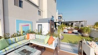 This expansive rooftop courtyard is a thoughtfully designed outdoor retreat that blends contemporary style with lush, resort-inspired landscaping. The space is organized into several distinct zones, each offering a unique experience. A wide wraparound deck finished in warm composite wood planks serves as the primary gathering area, furnished with colorful striped sectional sofas, accent pillows, and a central fire pit table — perfect for socializing against a backdrop of panoramic city views framed by sleek glass railings.

A shaded pergola zone provides a cooler sanctuary, featuring lounge chairs arranged before a living green wall where climbing vines cascade across a trellis structure. Nearby, a dedicated alfresco dining area offers a large mosaic-topped table with ample seating beneath the pergola, ideal for outdoor meals. A built-in outdoor kitchen and grill station supports the entertaining function of the space.

An open lawn area with artificial turf, tall potted palms, and ornamental plantings adds a grounding, park-like element. Bold contemporary wall art punctuates the architecture, while a sculptural geometric pendant light anchors the pergola's interior. A striped concrete walkway connects the various zones and leads through curated succulent garden beds, tying the entire courtyard together with a cohesive, design-forward aesthetic.