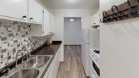 This single-floor unit offers a functional and straightforward layout across several well-defined spaces. The living area is finished with neutral wall-to-wall carpet and crisp white walls, complemented by a large window fitted with a window-mounted air conditioning unit, allowing natural light to fill the room. A baseboard heating system runs along the perimeter, providing an efficient heating solution throughout the space.

The galley-style kitchen is efficiently designed, featuring white cabinetry both above and below the counter, dark granite-look countertops, a mosaic tile backsplash, a double basin sink, and white appliances including a gas range and refrigerator. Wood-look flooring runs through the kitchen and extends into adjoining areas, adding warmth to the space. A ceiling fan overhead provides additional air circulation.

A hallway connects the main living area to additional rooms, including a carpeted bedroom with a window fitted with vertical blinds and baseboard heating, as well as a second bedroom or flex space with hardwood-style flooring. The bathroom features large-format white wall tiles, a soaking tub, a pedestal-style vanity with a mirror, and clean white fixtures throughout, creating a bright and functional environment.