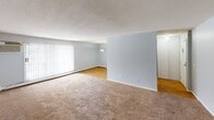 This single-floor residential unit offers a practical and well-organized layout across several distinct living areas. The open living and dining space is generously sized, featuring neutral-toned carpet, light blue-gray walls, and large vertical-blind-covered windows and a sliding glass door that allow ample natural light to fill the room. A baseboard heating system runs along the exterior walls, and a wall-mounted air conditioning unit provides climate control. The dining area transitions to hardwood-style flooring, anchored by a pendant light fixture overhead.

The galley-style kitchen is efficiently designed with white cabinetry, a double-basin sink, laminate countertops, and white appliances including a gas range and refrigerator. The hardwood-style flooring continues here, and a door provides direct exterior access.

The bedrooms feature wall-to-wall carpet, bright white walls, natural light from windows with blinds, baseboard heating, and flush-mount ceiling light fixtures. One bedroom also includes a wall-mounted air conditioning unit.

The bathroom is finished with clean white square tile surrounding a full-sized soaking tub with an overhead showerhead and chrome fixtures, complemented by soft gray walls. Textured ceilings are consistent throughout the unit, contributing to the space's classic residential character.