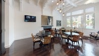 This single-story clubhouse delivers a refined, upscale atmosphere designed for community gatherings and relaxed socializing. The main lounge area commands attention with its soaring ceilings accented by decorative exposed beams and crisp white walls that amplify the sense of openness. Rich, dark hardwood flooring runs throughout, providing a warm contrast to the bright, airy interior.

The seating area is thoughtfully arranged with round dark tables surrounded by woven rattan and wood-framed chairs, creating an inviting, lounge-like setting. A fireplace serves as a focal point, flanked by curated artwork and a wall-mounted display screen. Statement pendant light fixtures featuring globe-style glass shades hang overhead, adding a modern, artistic touch.

Expansive windows flood the space with natural light while offering direct views of the outdoor pool and surrounding landscaping, seamlessly connecting the interior to the exterior environment.

The wet bar area is equally impressive, finished with dramatic dark cabinetry, bold veined stone countertops, a built-in wine rack with a distinctive X-pattern design, and an ornamental metalwork backsplash featuring an elegant tree motif. Globe pendant lights illuminate this functional yet visually striking entertaining space.