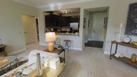 This single-floor residence offers a thoughtfully designed open-concept layout that promotes a comfortable and functional living experience. The living area is anchored by plush wall-to-wall carpeting and features elegant crown molding that adds a refined architectural touch to the space. A tufted sofa and coffee table arrangement highlights the generous proportions of the living room, which flows naturally toward the kitchen and entryway.

The kitchen is a standout feature, showcasing rich dark cabinetry that contrasts beautifully with light granite countertops and a classic tile backsplash. Stainless steel appliances, including a refrigerator, contribute to the kitchen's modern and polished aesthetic. A raised breakfast bar with industrial-style bar stools creates an inviting transition between the kitchen and living area, making it ideal for casual dining or entertaining.

The entryway is defined by durable tile flooring, providing a practical and attractive transition zone as you enter the home. The neutral color palette throughout — soft grays, warm whites, and deep espresso tones — creates a cohesive and welcoming ambiance. Track lighting in the kitchen ensures ample illumination, while the overall layout maximizes the sense of openness and ease of movement throughout the space.