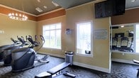 This fitness center is housed in a single-story space designed to support a range of workout activities. The room features warm beige and tan walls accented by a contrasting darker upper border, giving the space a defined, structured aesthetic. Crown molding adds a refined architectural touch throughout.

Natural light fills the room through multiple white-framed windows that offer views of the surrounding greenery, creating an open and inviting atmosphere. The patterned carpet flooring adds visual interest while providing a practical surface underfoot.

Equipment includes an elliptical machine and a weight bench with free weights, catering to both cardio and strength training needs. A flat-screen television is mounted high on the wall, allowing users to stay entertained during workouts. A sanitation station is conveniently positioned for equipment cleaning, reflecting a commitment to hygiene and user comfort.

Fitness center policy guidelines are posted prominently on the wall, ensuring users are informed of operating hours and usage rules. Recessed ceiling lighting supplements the natural light, keeping the space bright and functional throughout the day.