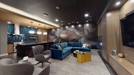 This thoughtfully designed single-floor amenity space combines elegance and functionality across several distinct zones. The centerpiece of the lounge area is a sweeping, floor-to-ceiling abstract mural in deep earth tones — burgundy, slate, and cream — that commands immediate attention and sets a dramatic, artistic tone throughout. A plush teal velvet sectional sofa anchors the seating area, complemented by sculptural gray swivel chairs and a geometric gold coffee table, all arranged atop a soft area rug. Opposite the mural, a full-width warm wood media wall houses a large flat-screen television and a sleek linear gas fireplace below, creating an inviting focal point for relaxed gatherings. Recessed ceiling lighting throughout provides a layered, ambient glow against the dark ceiling finish. The catering kitchen is equally impressive, featuring herringbone deep-blue tile backsplash, warm wood cabinetry, open floating shelves, stainless steel professional-grade appliances, a dark island with sink, and elegant pendant lighting. Adjacent to the lounge, a private conference room accommodates a large group around a long table set with leather chairs, illuminated by a sculptural multi-globe chandelier and equipped with a wall-mounted display screen. Dark hardwood-style flooring ties the zones together, creating a cohesive, upscale ambiance throughout.