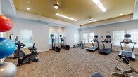 This single-floor fitness center offers a well-equipped workout environment designed to support a variety of exercise routines. The room is architecturally distinguished by a tray ceiling finished in a warm gold tone, which contrasts with the light gray walls below and adds visual interest to the space. Recessed lighting and flush-mount ceiling fixtures provide consistent, bright illumination throughout.

Natural light enters through multiple windows fitted with white horizontal blinds, positioned along several walls to create an open, airy atmosphere. The flooring consists of durable, textured carpet tile in a neutral earth-tone pattern, well-suited for a fitness environment.

Cardio equipment is well-represented, with multiple treadmills, elliptical trainers, an upright stationary bike, a recumbent bike, and a rowing machine distributed throughout the room. Each cardio machine is equipped with an individual monitor or tablet mount. Strength and flexibility training is supported by a weight bench, an abdominal craning bench, and a dedicated nook housing a yoga mat and a tiered rack of stability balls in assorted sizes.

Sanitation stations are mounted on the walls, and the space includes at least one accessible entry door. The layout provides ample circulation space between equipment, contributing to a functional and comfortable workout experience.
