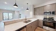 Located in downtown Winona, Minnesota, this thoughtfully designed 2-bedroom, 1-bathroom unit offers a well-appointed living experience with modern finishes throughout. The open-concept kitchen and living area serves as the heart of the home, where warm hardwood-style flooring and large black-framed windows fill the space with natural light. The kitchen is outfitted with taupe shaker-style cabinetry, quartz countertops, a white tile backsplash, stainless steel appliances, and a double-basin sink, all illuminated by stylish pendant lighting. Sliding glass doors lead to a private balcony with sweeping views of the Mississippi River, the surrounding bluffs, and the iconic Winona bridge. Both bedrooms feature plush carpeting, ceiling fans, and large windows overlooking the downtown cityscape. A well-lit hallway connects the bedrooms to the full bathroom, which is finished with tile flooring and marble-style wall accents. Three closets provide ample storage, while a dedicated laundry room with a full-size washer and dryer adds everyday convenience. Clean lines, neutral tones, and quality materials define the overall aesthetic of this modern urban residence.