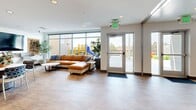 Located at 12444 Albrook Drive in Denver, Colorado, this thoughtfully designed community space offers a welcoming, open-plan layout across a single floor. The interior is unified by warm wood-look flooring, crisp white walls, and recessed lighting throughout, creating a bright and contemporary atmosphere.

The kitchen is a standout feature, showcasing rich dark wood cabinetry, white subway tile backsplash, and a large center island with a light quartz countertop. Seating along the island is provided by colorful patterned bar stools, while a pair of elegant gold loop pendant lights add a modern, upscale touch. The kitchen is fully equipped with built-in appliances, including a microwave and range.

The dining area is highlighted by a marble-topped table surrounded by eclectic patterned chairs, and the walls are adorned with a striking collection of vibrant abstract paintings that infuse the space with color and artistic energy. An additional long wooden work table provides flexible seating for larger gatherings.

The living room area is anchored by a generous caramel leather sectional sofa and a sleek white round coffee table. Floor-to-ceiling windows and a glass door flood the space with natural light and offer direct views of an outdoor playground, making it an inviting spot for relaxation and community interaction.