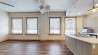 This single-floor unit showcases a thoughtful blend of industrial character and contemporary design. The open living area is defined by rich hardwood-style flooring, exposed HVAC ductwork overhead, and a generous arrangement of windows fitted with blinds that flood the space with natural light while offering views of the surrounding urban streetscape. A ceiling fan adds a functional accent to the airy main area.

The kitchen stands as a central highlight, featuring a curved island with a light wood-grain finish and a clean white countertop. Light-toned cabinetry lines the walls, complemented by a subway tile backsplash, stainless steel appliances including a full-sized refrigerator, an integrated sink, and pendant lighting suspended beneath a softly arched ceiling detail that frames the space elegantly.

The bedroom areas offer a contrast in finishes: one room features the same hardwood-style flooring with crisp white walls, while another is finished with neutral carpeting and includes a closet with a sliding door. Each room benefits from a calm, understated palette. The 3D scan reveals a well-proportioned floor plan with clearly defined zones, reinforcing the unit's practical yet stylish layout throughout.