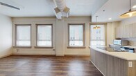 This single-floor unit showcases a thoughtful blend of industrial character and contemporary design. The open living area is defined by rich hardwood-style flooring, exposed HVAC ductwork overhead, and a generous arrangement of windows fitted with blinds that flood the space with natural light while offering views of the surrounding urban streetscape. A ceiling fan adds a functional accent to the airy main area.

The kitchen stands as a central highlight, featuring a curved island with a light wood-grain finish and a clean white countertop. Light-toned cabinetry lines the walls, complemented by a subway tile backsplash, stainless steel appliances including a full-sized refrigerator, an integrated sink, and pendant lighting suspended beneath a softly arched ceiling detail that frames the space elegantly.

The bedroom areas offer a contrast in finishes: one room features the same hardwood-style flooring with crisp white walls, while another is finished with neutral carpeting and includes a closet with a sliding door. Each room benefits from a calm, understated palette. The 3D scan reveals a well-proportioned floor plan with clearly defined zones, reinforcing the unit's practical yet stylish layout throughout.