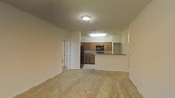 This single-floor residence offers a clean, open-concept layout that connects the living area and kitchen in a practical and cohesive design. The living area is finished with neutral-toned wall-to-wall carpeting, creating a warm and comfortable atmosphere throughout the space. Smooth, light-colored walls and flush ceiling lighting contribute to a bright and airy feel.

The kitchen is positioned at the rear of the main living area and features warm wood-toned cabinetry that provides ample storage. It is equipped with black appliances, including a refrigerator, range with oven, and over-the-range microwave. A raised breakfast bar counter serves as both a functional workspace and a natural divider between the kitchen and living areas, allowing for easy interaction between the two spaces.

A doorway visible from the main living area leads to additional rooms, suggesting a well-organized floor plan with private spaces set apart from the common living areas. The consistent neutral color palette throughout the space creates a cohesive, move-in-ready aesthetic. Overall, the layout is designed for efficiency and ease of living, making it a straightforward and functional single-level home.