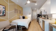 This thoughtfully designed studio unit makes efficient use of its open-plan layout, seamlessly integrating living, sleeping, dining, and cooking areas into a single cohesive space. The interior is anchored by a striking gold and white geometric accent wall that adds warmth and a modern artistic flair to the sleeping area, complemented by curated wall décor and a platform bed with under-bed storage. Light-toned hardwood-style flooring runs throughout, enhancing the bright and airy atmosphere created by recessed lighting and a ceiling fan overhead. The galley-style kitchen lines one wall and is outfitted with stainless steel appliances including a refrigerator, microwave, and range, alongside white shaker cabinetry with matte black hardware, a subway tile backsplash, and a clean white countertop with an integrated sink. A built-in wooden desk with a large mounted television above serves as a functional workspace and entertainment area. A compact dining nook with a round table and seating provides a defined area for meals. The private bathroom features wood-look tile flooring, a walk-in shower with a sliding glass door and black fixtures, a vanity, and clean white wall tiling, completing this efficiently appointed studio.