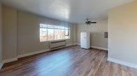 This thoughtfully designed single-floor studio offers a clean, open layout finished with light-toned hardwood flooring throughout, creating a warm and cohesive aesthetic. The main living area is airy and well-lit, anchored by a wide picture window fitted with horizontal blinds that frame a striking urban skyline view, with city buildings and tree canopies visible beyond. A ceiling fan with a brushed nickel finish provides both function and a subtle decorative touch overhead. A wall-mounted radiant heating unit runs beneath the window, ensuring year-round comfort. Adding a distinctive retro character to the space, a cream-colored SMEG refrigerator stands as a design focal point. The compact kitchen is neatly tucked into a dedicated alcove, featuring white shaker-style cabinetry extending to the ceiling for maximized storage, a stainless steel sink, a neutral countertop, and a black gas range. An electrical panel is conveniently recessed into the adjacent wall. The neutral, warm-toned walls throughout complement the light flooring and contribute to the space's bright, inviting ambiance. The open floor plan allows for flexible furniture arrangements, making this a versatile and functional urban living space.