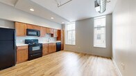 Located at 2828 Euclid Ave in Cleveland's Midtown corridor, this one-bedroom, one-bathroom unit blends urban industrial character with modern finishes. The open-plan living and kitchen area is anchored by light hardwood flooring, recessed lighting, and tall windows that draw in generous natural light. The kitchen is well-appointed with warm wood cabinetry, a full suite of black appliances including a refrigerator, range, microwave, and dishwasher, and a clean white countertop with a stainless steel sink. Exposed spiral ductwork running along the ceiling reinforces the space's industrial loft aesthetic. A long hallway connects the main living area to the private bedroom, which features plush carpeting and a striking rustic barn door crafted from reclaimed wood on a black sliding track. The bedroom also includes built-in shelving for additional storage, along with a ceiling fan. The bathroom continues the industrial theme with a full exposed brick accent wall, wood-look tile flooring, a walk-in shower, and a white vanity. In-unit laundry is also accessible from the bedroom area, adding everyday convenience to this thoughtfully designed urban residence.