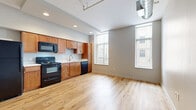 Located at 2828 Euclid Ave in Cleveland's Midtown corridor, this one-bedroom, one-bathroom unit blends urban industrial character with modern finishes. The open-plan living and kitchen area is anchored by light hardwood flooring, recessed lighting, and tall windows that draw in generous natural light. The kitchen is well-appointed with warm wood cabinetry, a full suite of black appliances including a refrigerator, range, microwave, and dishwasher, and a clean white countertop with a stainless steel sink. Exposed spiral ductwork running along the ceiling reinforces the space's industrial loft aesthetic. A long hallway connects the main living area to the private bedroom, which features plush carpeting and a striking rustic barn door crafted from reclaimed wood on a black sliding track. The bedroom also includes built-in shelving for additional storage, along with a ceiling fan. The bathroom continues the industrial theme with a full exposed brick accent wall, wood-look tile flooring, a walk-in shower, and a white vanity. In-unit laundry is also accessible from the bedroom area, adding everyday convenience to this thoughtfully designed urban residence.