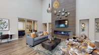 This well-appointed single-floor space is designed to impress with its striking architectural details and contemporary interior design. The central lounge area commands attention with dramatically soaring ceilings that amplify the sense of openness and grandeur. The focal point of the room is a full-height, horizontally-striped tile wall housing a wall-mounted flat-screen television above a sleek, linear electric fireplace with vivid blue flame accents. Decorative stone-like sculptures are artfully arranged on the tile surround, adding an organic, sculptural element to the space. The lounge is furnished with a plush gray sectional sofa accented by warm amber throw pillows, set atop a layered area rug in rich jewel tones. Woven rattan pendant lights cascade from the ceiling, lending a warm, textural glow to the space. Wood-look flooring runs throughout, grounding the design with a natural warmth. An adjacent area features a long wall-mounted work surface with leather chairs beneath a pair of bold circular abstract art pieces. A separate kitchenette area showcases teal cabinetry with matte black hardware, white quartz countertops, a textured scallop-pattern tile backsplash, and a utility sink — all illuminated by a vintage-style pendant light. Natural light filters in through a nearby window dressed in patterned drapery, completing the inviting and functional atmosphere.