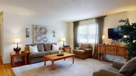 This single-floor home offers a thoughtfully designed layout with warm, inviting interiors throughout. The living room serves as the heart of the home, showcasing rich hardwood floors, a large area rug, and generous natural light streaming through windows dressed with elegant gray drape curtains. The space is anchored by a solid pine coffee table and complemented by tasteful wall art, creating a relaxed yet polished atmosphere.

Adjacent to the living area, the dining space features the same hardwood flooring, a ceiling fan with light fixture, and large windows that bathe the room in natural light. A side door provides convenient outdoor access directly from the dining area.

The kitchen is equipped with warm oak cabinetry, ample counter space, a gas range with overhead ventilation, and a dishwasher, offering a functional setup for everyday cooking.

The bedrooms are comfortable and well-lit, each featuring hardwood or carpeted floors, natural light from sizable windows with blinds and draping curtains, and adequate space for furnishings. One bedroom includes a dedicated workspace with a mission-style desk and chair.

The bathroom includes a glass-enclosed shower stall with classic white tile surround, maintaining a clean and practical aesthetic throughout. Overall, the home presents a cohesive, warm design with consistent natural wood tones and neutral color palettes.