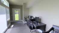 This single-floor space is designed to serve as a practical and comfortable work environment. The office area is anchored by an L-shaped dark-finish desk that provides ample workspace, accommodating dual computer workstations complete with monitors, a keyboard, and a printer. Two ergonomic mesh-back chairs on casters offer flexible seating arrangements suited for productivity. The desk unit includes integrated storage cabinets below, providing functional organization solutions.

The room features neutral, light-toned walls that create a clean and professional atmosphere, complemented by wall-to-wall carpeting in a soft gray tone that adds warmth underfoot. The angled ceiling line adds a subtle architectural character to the space.

Natural light is a notable feature, with a white-framed glass-panel door at the far end of the room drawing in daylight and framing a view of a verdant green lawn and surrounding trees beyond. This connection to the outdoors enhances the brightness and openness of the workspace, making it an inviting and well-lit environment suited for daily office use.