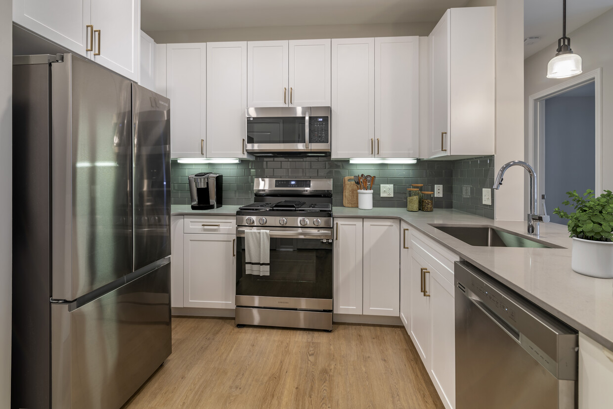 Renovated Package II kitchen with stainless steel appliances, grey quartz countertops, white cabinetry, grey tile backsplash, and hard surface flooring - Avalon at Assembly Row