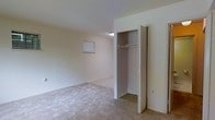 This single-story space includes a bedroom that showcases a clean, straightforward layout with neutral-toned walls and light-colored carpeting throughout. A built-in closet with bifold doors is positioned along one wall, offering a full-height hanging rod and overhead shelf for organized storage. The interior lighting fixture inside the closet ensures visibility within the storage area. A small window on the far wall allows a modest amount of natural light into the room, complementing the bright, airy feel created by the white ceiling and pale wall tones. The overall ambiance is simple and understated, presenting a blank canvas well-suited for personalization. The neutral color palette and carpeted flooring contribute to a quiet, comfortable atmosphere throughout the space.