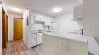 This single-floor unit offers a well-organized and functional layout across several distinct living areas. The kitchen is a standout feature, finished with crisp white cabinetry, light countertops, and white appliances including a refrigerator, dishwasher, and electric range. A ventilation hood and under-cabinet storage maximize utility, while wood-look luxury vinyl plank flooring ties the space together. A peninsula-style counter provides a natural divide between the kitchen and the rest of the unit. The living area is generously proportioned, bathed in natural light through a large sliding glass door that opens to a balcony, offering views of lush greenery outside. A wall-mounted air conditioning unit and baseboard heating provide year-round climate control. The bedroom is carpeted for comfort, offering a quiet and cozy retreat with a closet and a ceiling-mounted light fixture. The bathroom is clean and practical, equipped with a full-size soaking tub and shower combination, a toilet, and wood-look flooring that complements the rest of the unit. Throughout the space, textured ceilings, neutral wall tones, and consistent flooring create a cohesive and move-in-ready environment.