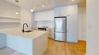 Located at 1250 Avenue Thérèse-Lavoie-Roux in Montréal's Outremont neighbourhood, this well-appointed two-bedroom, two-bathroom unit offers a thoughtfully designed single-floor layout. The modern kitchen serves as a central highlight, featuring a large island with a white quartz countertop, matte black faucet, floor-to-ceiling white cabinetry, a white tile backsplash, stainless steel appliances including a full-size refrigerator and range, and warm hardwood flooring. Pendant lighting adds a contemporary touch overhead. Both bedrooms are bright and minimalist, benefiting from large windows that bring in natural light and offer views of the surrounding urban streetscape. The exposed concrete ceiling adds an industrial character that contrasts elegantly with the clean white walls and light hardwood floors throughout. Each bedroom is served by its own dedicated bathroom: one features a sleek glass-enclosed walk-in shower with large-format tiles and a warm wood-toned vanity, while the second includes a soaker tub with a glass partition and coordinating fixtures. Two closets provide practical storage, and an in-unit washer and dryer add everyday convenience. The overall aesthetic is clean, modern, and carefully finished.
