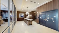 Located at 1900 Chapman Ave in Rockville, Maryland, this thoughtfully designed lobby level sets a polished tone for the building it serves. The main entry area showcases large-format cream tile flooring, warm wood-paneled accent walls, and a striking crystal chandelier suspended above a gold-framed communal table with upholstered stools — creating an inviting gathering space bathed in ambient light. Floor-to-ceiling glass partitions along one side offer views into an adjacent lounge or conference area, enhancing the sense of openness and connectivity. A built-in bench with a rich red cushion adds a functional yet stylish touch to the seating arrangements. The mail center is a standout feature, housing an extensive array of individual resident mailboxes set within warm wood cabinetry, complemented by abstract artwork that adds character to the space. Nearby, a bank of dark, geometrically patterned package lockers provides secure parcel storage. Connecting corridors are finished with coordinating tile flooring, wainscoting in muted grey tones, and sculptural ceiling-mounted light fixtures, maintaining a cohesive design language throughout. The overall ambiance is modern and sophisticated, balancing practicality with refined aesthetic detail.
