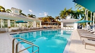 Located at 8485 Hammocks Boulevard in Miami, FL, this outdoor amenity area showcases a generously sized resort-style swimming pool as its centerpiece. The crystal-clear, blue pool is framed by a wide, paver-tiled sundeck furnished with rows of lounge chairs and teal market umbrellas, providing ample space for relaxation under the South Florida sun. Towering royal palms line the perimeter, creating a lush, tropical atmosphere that enhances the overall resort ambiance. A well-appointed poolhouse anchors one end of the space, featuring large glass windows, a standing-seam metal roof with a decorative cupola, and an adjacent pergola-covered lounge area outfitted with outdoor seating—ideal for socializing or unwinding in the shade. A separate pool rules structure and what appears to be a pool bar or service station adds further functionality to the space. The surrounding landscaping incorporates manicured planters and greenery that complement the clean, white architectural finishes of the buildings visible in the background. The overall design reflects a polished, tropical aesthetic consistent with Miami's warm climate and outdoor lifestyle, offering residents a well-equipped and visually striking communal outdoor retreat.