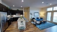 This single-story space showcases an open-concept layout that integrates the kitchen and living area into one cohesive, flowing environment. The kitchen is outfitted with dark espresso cabinetry, a stone tile backsplash, and granite countertops that extend across a central island with seating. The island serves as both a prep area and casual dining space, featuring lower open shelving for added storage and display. Recessed lighting illuminates the kitchen, complementing the warm tones of the hardwood-style flooring that runs throughout the space. The transition into the living area is seamless, creating a generous sense of openness. The living space is anchored by a bold blue sofa, modern accent chairs, and a patterned area rug, all arranged around a sleek coffee table. Wall-mounted decorative accents and framed artwork add visual interest to the neutral walls. A tall floor lamp provides warm ambient lighting, contributing to the inviting atmosphere. A hallway visible in the background suggests additional rooms beyond the main living area. Overall, the space combines modern finishes with a warm, welcoming ambiance.
