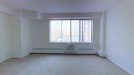 This single-floor space presents a clean, open layout finished with wall-to-wall carpeting and crisp white walls throughout. The main room is defined by a large picture window fitted with vertical blinds, allowing an abundance of natural light to fill the interior while offering views of the surrounding exterior environment, including neighboring structures and greenery. Beneath the window, a wall-mounted air conditioning unit is flanked by continuous baseboard heating units that run along the perimeter of the room, providing a comprehensive climate control system suitable for year-round comfort. The ceiling features a subtle dropped section framing the window wall, adding a minor architectural detail to the otherwise straightforward, minimalist design. The open floor plan provides flexibility for a variety of uses and configurations. The overall ambiance is bright, neutral, and functional, with the generous window serving as the focal point of the space and ensuring a well-lit environment throughout the day.