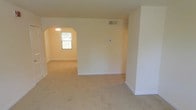 This single-floor space presents a clean, understated interior defined by neutral cream-toned walls and wall-to-wall beige carpeting that extends throughout the living areas. The most distinctive architectural feature is a rounded arched doorway that connects the main living area to an adjoining room, adding a classic design element to an otherwise straightforward layout. The secondary room beyond the arch is illuminated by a window that allows natural light to filter in, brightening the space. A ceiling light fixture is visible through the archway, providing additional overhead lighting. The main living area is generously proportioned, offering an open feel with minimal visual obstructions. White baseboards provide clean definition between the walls and flooring, contributing to the overall bright and tidy aesthetic. A ceiling vent indicates a central HVAC system, ensuring climate control throughout. The walls are smooth and neutral, offering a versatile backdrop for a variety of interior styles. Overall, the space feels light, open, and move-in ready, with timeless architectural details that lend a subtle sense of character.
