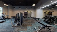 This single-floor fitness center offers a thoughtfully designed workout space that blends modern aesthetics with high-performance functionality. The interior features a striking dark ceiling with exposed structural elements and bold pendant lighting, contrasting elegantly against the crisp white walls and patterned teal-and-gray carpet tiles that define the open exercise floor.

The space is comprehensively equipped to support a wide variety of training styles. Strength training is well-represented with Matrix selectorized machines, adjustable benches, and a full rack of kettlebells and medicine balls on a dedicated storage stand. A wall-mounted stall bar provides additional functional training versatility. For combat and conditioning, a freestanding heavy punching bag adds a dynamic element to the floor layout.

Cardio options include Matrix treadmills positioned near large windows and a Jacob's Ladder climber for high-intensity interval training. A stair-style incline trainer further diversifies the cardio offerings. Full-length mirrors along one wall enhance the sense of space and support form-checking during workouts.

A built-in counter area with a flat-screen TV and beverage cooler provides a convenient amenity station. Large glass double doors and generous windows along one wall invite natural light and offer views of the surrounding outdoor courtyard, creating a bright and energizing training environment.