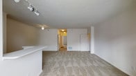 This single-floor residential space offers a practical and functional layout designed for comfortable everyday living. The main living area is generously sized, featuring neutral wall-to-wall carpeting that adds warmth and a soft underfoot feel throughout the space. The textured ceiling adds subtle visual interest overhead, while a ceiling fan provides both air circulation and ambient lighting. Track lighting is also present, offering flexible and directed illumination across the room. A partial countertop or bar-height divider is visible along one side, hinting at a kitchen or dining area just beyond. The open layout creates a seamless flow between the living space and the connecting rooms, which are accessible through a hallway visible in the background. A closet with a white panel door is positioned along one wall, providing convenient storage. Floor vents indicate a forced-air climate control system throughout. The clean white walls and bright overhead lighting give the space a fresh, move-in-ready feel. Overall, the layout is efficient and well-connected, making it a versatile and practical living environment.