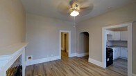 This single-floor residence combines classic architectural charm with a clean, neutral aesthetic. The main living area showcases warm hardwood-style flooring that runs throughout, complemented by crisp white baseboards that frame each room with a polished finish. A ceiling fan with a light fixture is centrally mounted, providing both ventilation and ambient lighting to the space.

A standout architectural detail is the elegant arched alcove built into the wall, adding a timeless character to the interior and offering a decorative niche that breaks up the clean lines of the room. A traditional rectangular doorway with white trim leads into a hallway, connecting the living area to additional rooms beyond. The hallway, visible through the doorway, continues the same warm flooring, suggesting a cohesive design throughout the home.

The walls are finished in a soft, warm greige tone that pairs naturally with the flooring, creating a calming and inviting ambiance. The open and uncluttered layout of the main room allows for flexible furnishing arrangements. Overall, the space reflects a well-maintained interior with thoughtful architectural touches that blend traditional design elements with a simple, functional layout suited for comfortable everyday living.