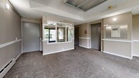 Located at 14921 104th Ave in Surrey, BC, this single-floor common area serves as the main lobby and entry point for the building. The space is thoughtfully designed to facilitate smooth resident and visitor flow, incorporating key functional elements throughout.

The lobby is finished with patterned, multi-tone carpeting that adds texture underfoot, while the walls are painted in a neutral warm beige with a crisp white chair rail running along the perimeter, lending a clean and orderly appearance. A textured ceiling with recessed panel detailing adds a subtle architectural character overhead.

A built-in reception or mailbox window with a white-framed pass-through is a central feature, offering a practical point of interaction. The elevator, framed by grey metal doors, provides vertical access to the rest of the building, and is positioned alongside a secondary door, ensuring efficient circulation.

Large glass entry doors at the front of the lobby welcome natural light into the space and offer views of the lush greenery outside, contributing to a bright and inviting atmosphere. A baseboard heating unit runs along the exterior wall, ensuring comfort year-round. Safety and building management fixtures, including fire safety equipment and an intercom panel, are clearly integrated throughout. The overall ambiance is functional, tidy, and well-maintained, appropriate for a residential building lobby.
