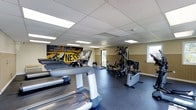 This fitness center offers a comprehensive workout environment designed to accommodate a range of exercise needs. The space is finished with a durable dark rubber floor, ideal for high-traffic athletic use, and features a drop-panel ceiling with recessed fluorescent lighting that keeps the area evenly and brightly lit throughout.

Along one side of the room, a row of treadmills provides ample cardio options, while a multi-station strength training machine occupies the opposite area, offering a versatile range of resistance exercises. Additional cardio and conditioning equipment can be found throughout the floor, ensuring a well-rounded selection of fitness tools.

Natural light filters in through windows along the walls, contributing to an open and energizing atmosphere. The wainscoted walls feature a warm neutral tone, complemented by a bold, black-and-yellow motivational graphic mural that serves as a focal point and adds an energetic, gym-like ambiance to the space.

Overall, the fitness center is thoughtfully laid out to maximize usable floor space while maintaining clear pathways between equipment stations, making it functional and accessible for a variety of fitness routines.