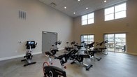 Located at 1 Mirada Dr N in Lewis Center, Ohio, this modern fitness facility offers a well-appointed, single-floor layout designed to support an active lifestyle. The centerpiece of the space is a generously sized cycling studio that immediately impresses with its soaring double-height ceilings and recessed lighting, creating a bright and energizing atmosphere. Large windows positioned at an elevated level allow natural light to pour into the room throughout the day, enhancing the open and airy feel of the interior. The studio is outfitted with a full lineup of professional-grade LifeCycle stationary bikes, each equipped with integrated display screens, arranged to accommodate group cycling classes or individual workouts. The flooring features a light-toned wood-look surface with a subtle herringbone pattern, adding a refined aesthetic touch to the functional space. Neutral wall tones and a clean, minimalist design keep the focus on performance and movement. A set of double doors provides convenient access, connecting the cycling area to the broader facility. The overall ambiance is polished and contemporary, reflecting a thoughtfully designed environment built to deliver an effective and motivating fitness experience.