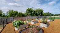 Located at 12302 S Yukon Ave in Glenpool, Oklahoma, this thoughtfully designed community garden offers a dedicated outdoor growing space for residents of the surrounding multi-story residential community. The garden is fully enclosed, combining a wooden privacy fence along the perimeter with chain-link fencing and a gated entry, providing both security and a defined sense of place.

The garden features numerous galvanized steel raised planting beds of varying shapes and sizes, including rectangular and oval configurations, arranged in an orderly layout across a gravel ground surface that promotes drainage and ease of movement between beds. The raised beds are actively cultivated with a diverse mix of vegetables, herbs, and flowering plants, reflecting a vibrant and productive growing environment.

The gravel pathways between the beds allow easy access to all planting areas, while the open sky above ensures ample sunlight throughout the day. Mature trees visible beyond the fence line provide a natural backdrop and partial wind protection. The surrounding green lawn areas and residential buildings are visible from within the garden, reinforcing its role as a community-centered amenity. Overall, the space offers a functional, well-maintained, and inviting environment for gardening enthusiasts.