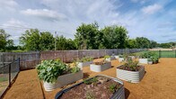 Located at 12302 S Yukon Ave in Glenpool, Oklahoma, this thoughtfully designed community garden offers a dedicated outdoor growing space for residents of the surrounding multi-story residential community. The garden is fully enclosed, combining a wooden privacy fence along the perimeter with chain-link fencing and a gated entry, providing both security and a defined sense of place.

The garden features numerous galvanized steel raised planting beds of varying shapes and sizes, including rectangular and oval configurations, arranged in an orderly layout across a gravel ground surface that promotes drainage and ease of movement between beds. The raised beds are actively cultivated with a diverse mix of vegetables, herbs, and flowering plants, reflecting a vibrant and productive growing environment.

The gravel pathways between the beds allow easy access to all planting areas, while the open sky above ensures ample sunlight throughout the day. Mature trees visible beyond the fence line provide a natural backdrop and partial wind protection. The surrounding green lawn areas and residential buildings are visible from within the garden, reinforcing its role as a community-centered amenity. Overall, the space offers a functional, well-maintained, and inviting environment for gardening enthusiasts.