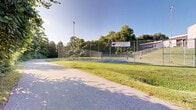 Located at 831 Cleveland St in Greenville, South Carolina, this single-level outdoor space offers a welcoming, park-like environment characterized by an abundance of mature trees, dense shrubs, and lush vegetation. A wide paved pathway winds through the grounds, providing a smooth and accessible route that invites leisurely walks amid the natural surroundings. Along the edges of the path, vibrant greenery thrives, including climbing vines, leafy shrubs, and a variety of native plants that create a rich, layered landscape. Sunlight filters through the tree canopy, casting a warm, dappled glow across the grounds throughout the day. A well-maintained tennis court, enclosed by chain-link fencing and equipped with overhead lighting, serves as a standout recreational feature of the property. The courts are set against a backdrop of open green lawn and mature trees, enhancing the overall sense of open-air recreation. The surrounding landscape is verdant and well-established, offering a sense of natural seclusion while remaining accessible. Nearby multi-story buildings are visible in the background, placing this outdoor space within a broader residential and recreational context in the heart of Greenville.