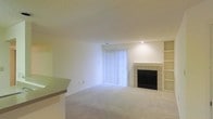 This single-floor space centers around a bright, open living area finished with neutral wall-to-wall carpeting and crisp white walls that create a clean, airy atmosphere. A tile-surround fireplace serves as the focal point of the main living area, flanked by built-in shelving and display niches that offer both functional storage and visual interest. Recessed lighting above the fireplace provides targeted illumination, complementing the natural light that filters into the room through a full-width sliding glass door dressed with vertical blinds. The sliding door not only brightens the interior but also provides access to an outdoor area. A partial countertop or bar overhang is visible along one edge of the space, suggesting an adjacent kitchen or dining area that transitions smoothly into the living room. The overall layout feels open and versatile, well-suited for a variety of furniture arrangements. The consistent use of light tones throughout — from the flooring to the walls and ceiling — contributes to a cohesive, understated aesthetic that emphasizes spaciousness and simplicity.