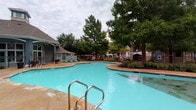 This outdoor pool area is a well-appointed recreational amenity designed for relaxation and leisure. The centerpiece is a generously sized, freeform swimming pool with crystal-clear turquoise water, accessible via stainless steel entry rails and steps. Adjacent to the main pool sits a spa, offering a dedicated space for unwinding. The expansive concrete sun deck surrounds the pool and is furnished with numerous lounge chairs and chaise lounges, providing ample space for sunbathing and socializing. Mature trees frame the perimeter of the area, offering natural shade and a sense of privacy, while manicured landscaping with shrubs and plantings adds color and texture throughout. A covered poolside structure with wood-trimmed doors serves as a pool house, providing convenient access to facilities and featuring large glass doors that overlook the pool. The area is enclosed by a wrought iron fence, ensuring safety and security. The overall ambiance is inviting and resort-like, with a well-maintained deck, vibrant greenery, and a layout that balances open sunlit spaces with shaded retreats — making it a versatile and appealing outdoor destination.