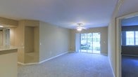 This single-story space centers around a spacious, open living area designed for comfort and functionality. The room features wall-to-wall carpeting in a neutral tone that complements the beige walls, creating a warm and cohesive aesthetic throughout. A ceiling fan with an integrated light fixture is mounted at the center of the smooth, flat ceiling, providing both air circulation and ambient lighting.

A defining feature of the living area is the full-width sliding glass door along the rear wall, which floods the space with natural light and offers direct views of lush outdoor greenery. The exterior view reveals well-maintained landscaping, enhancing the connection between the interior and the outdoors.

The walls are finished in a soft, neutral beige, providing a versatile backdrop suitable for a variety of décor styles. A recessed niche is visible along one wall, offering a built-in display or storage opportunity. White baseboards frame the perimeter of the room cleanly. The overall ambiance is calm and understated, with an open layout that allows for flexible furniture arrangements and a bright, airy feel supported by the generous natural light from the glass door.