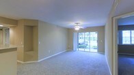 This single-story space centers around a spacious, open living area designed for comfort and functionality. The room features wall-to-wall carpeting in a neutral tone that complements the beige walls, creating a warm and cohesive aesthetic throughout. A ceiling fan with an integrated light fixture is mounted at the center of the smooth, flat ceiling, providing both air circulation and ambient lighting.

A defining feature of the living area is the full-width sliding glass door along the rear wall, which floods the space with natural light and offers direct views of lush outdoor greenery. The exterior view reveals well-maintained landscaping, enhancing the connection between the interior and the outdoors.

The walls are finished in a soft, neutral beige, providing a versatile backdrop suitable for a variety of décor styles. A recessed niche is visible along one wall, offering a built-in display or storage opportunity. White baseboards frame the perimeter of the room cleanly. The overall ambiance is calm and understated, with an open layout that allows for flexible furniture arrangements and a bright, airy feel supported by the generous natural light from the glass door.